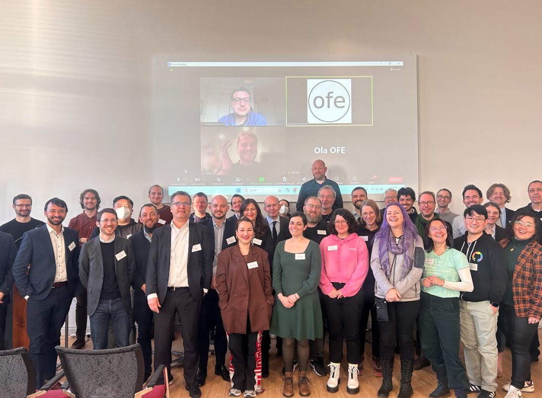 صورة جماعية للمشاركين في مؤتمر OFA. الكثير من الوجوه المبتسمة مع وجود الحاضرين الافتراضيين على الشاشة في الخلفية.